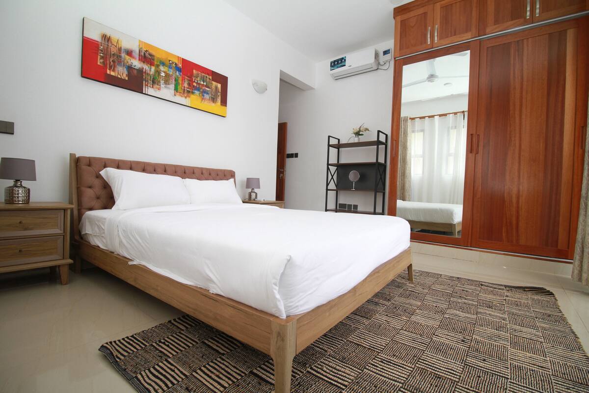 A bedroom with a wooden bed, a bedside table, a nightstand, a wall-mounted air conditioner, and a large wardrobe.