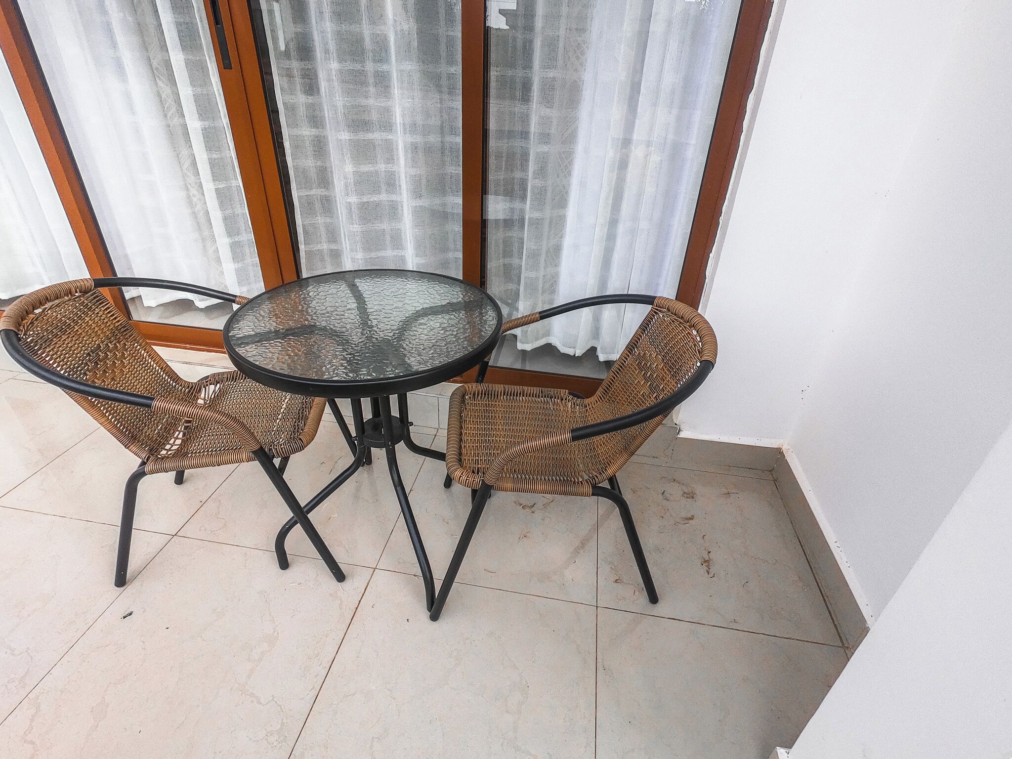 Una mesa con cubierta de vidrio y dos sillas de mimbre en una habitación con piso de baldosas y una ventana con cortinas.