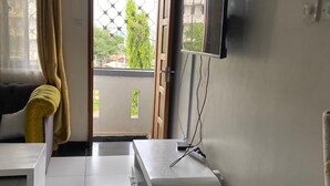 Une pièce avec un canapé blanc, une petite table blanche, un téléviseur fixé au mur et une fenêtre avec des rideaux.