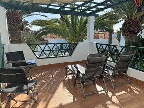 Een overdekt terras met stoelen en een tafel, met uitzicht op een zwembad en palmbomen.