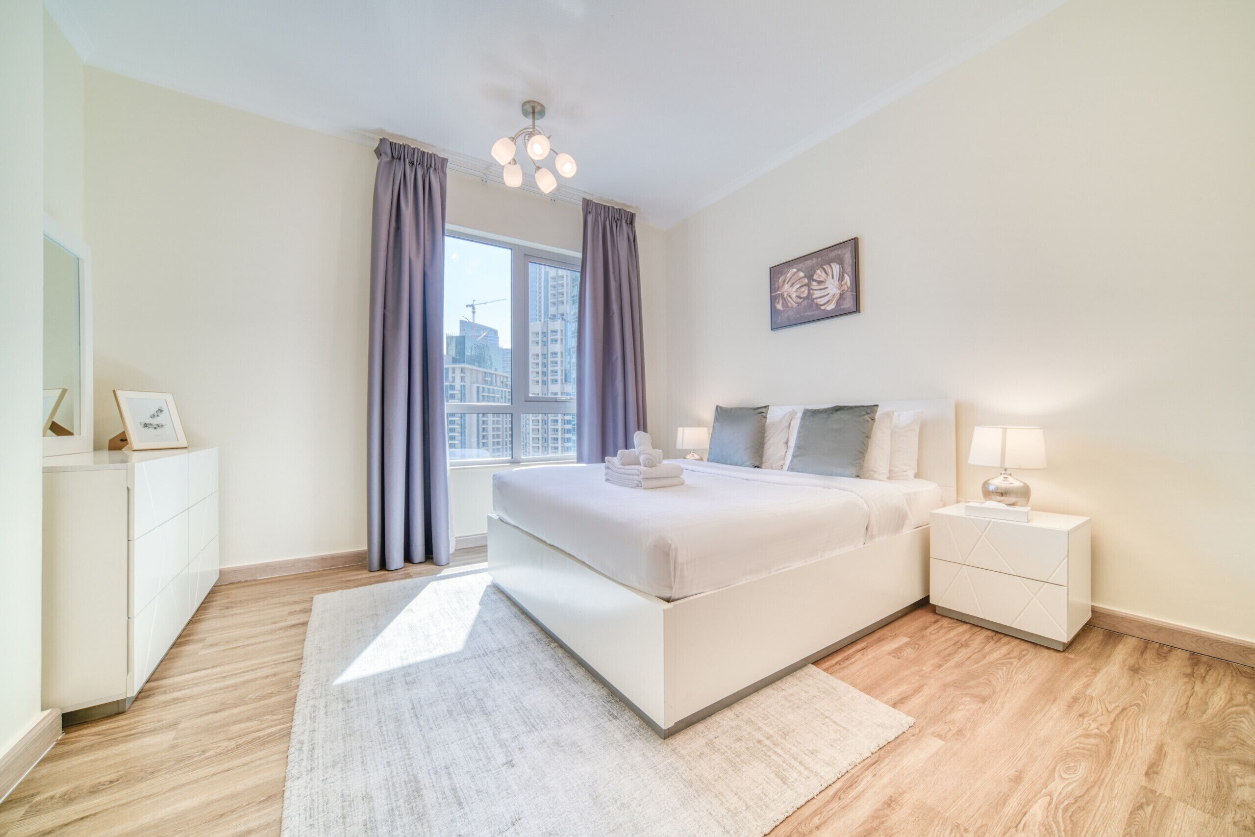 A modern bedroom with a large bed, bedside tables, a nightstand, a framed picture, and a view of the cityscape through the window.