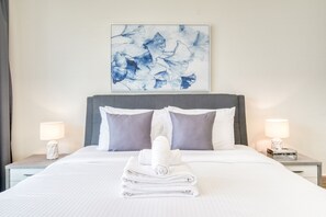 Een netjes opgemaakt bed met een opgevouwen handdoek, twee grijze kussens en een wit hoofdbord. Daarboven hangt een lijst met een abstract schilderij, en aan weerszijden van het bed staan nachtkastjes met lampen.