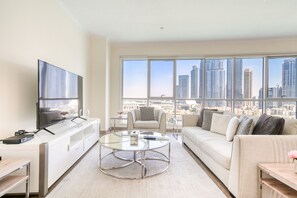 A modern living room with a flat-screen TV, a glass coffee table, a sofa with patterned cushions, and a view of a city skyline through large windows.