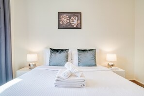 Una cama bien hecha con sábanas blancas y una toalla doblada, dos almohadas a juego, una obra de arte enmarcada sobre la cama y lámparas de noche en mesitas blancas.