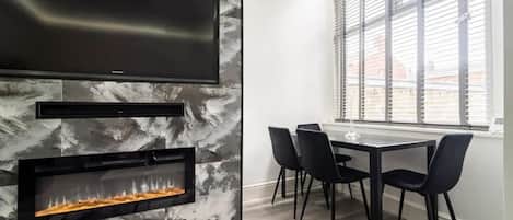 A modern dining area with a fireplace, a flat-screen TV, and a large window with blinds.