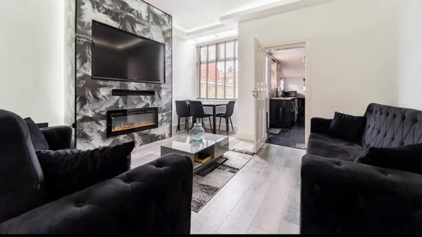 A modern living room with a fireplace, black sofas, a TV, and a dining area. - House With Modern Interior, Conveniently Located (Manchester)
