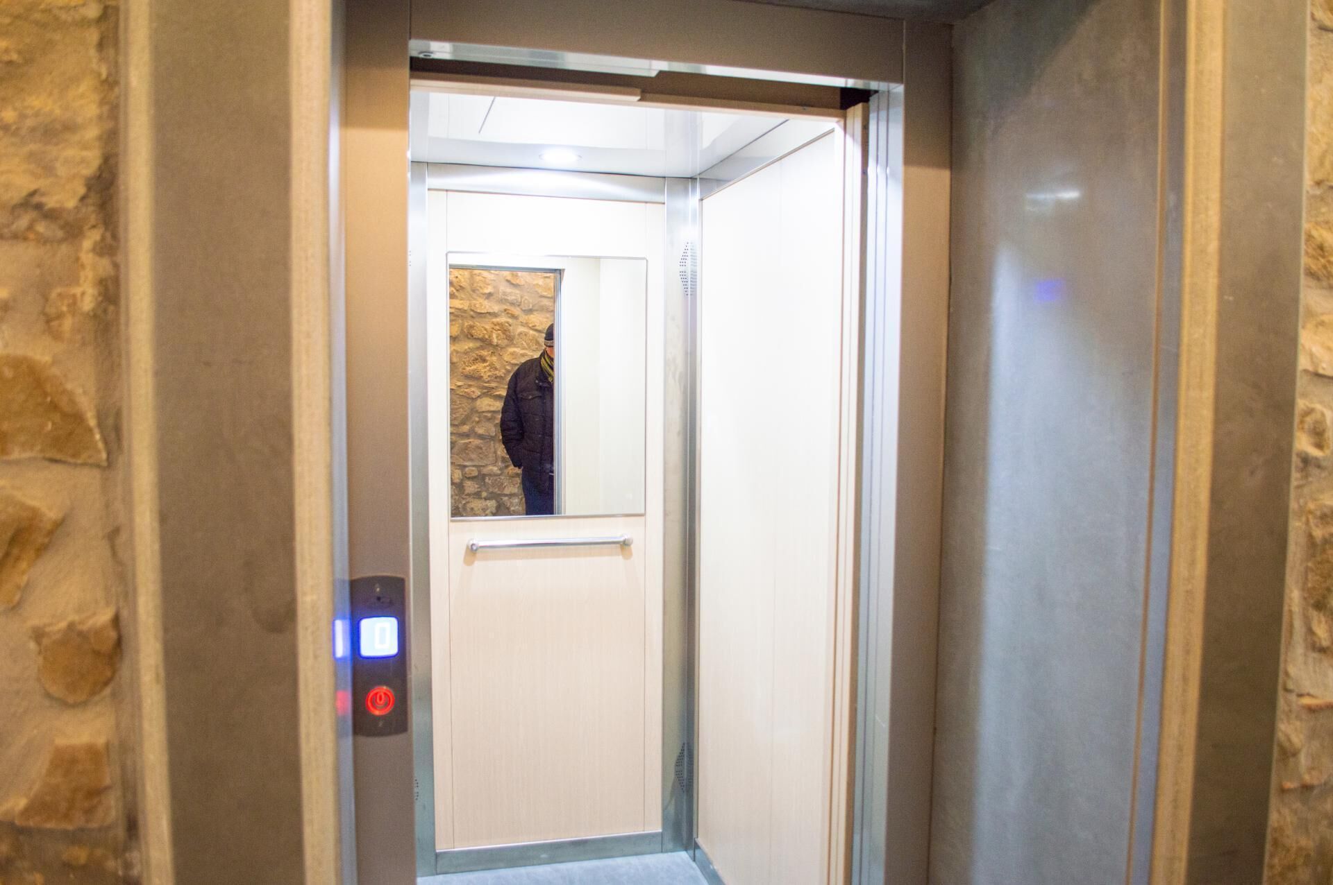 Interior de un ascensor con pared de piedra, una puerta y un panel de control con un botón rojo.
