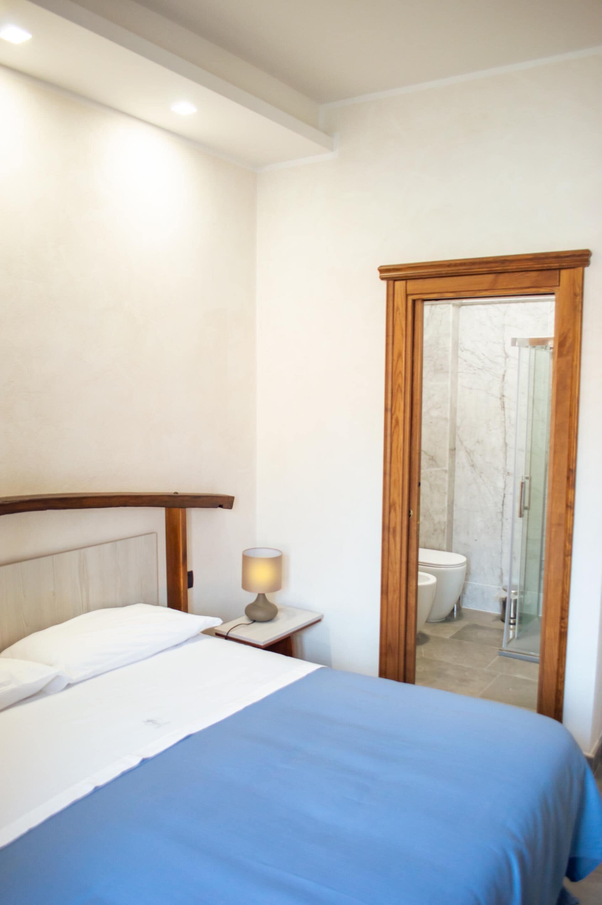 Une chambre à coucher avec un lit, une lampe de chevet et une porte en bois donnant sur une salle de bain équipée de toilettes et d’une douche.