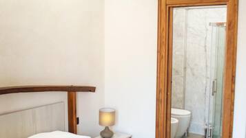 Un dormitorio con cama, una lámpara de noche, una puerta de madera que da a un baño con inodoro y ducha.