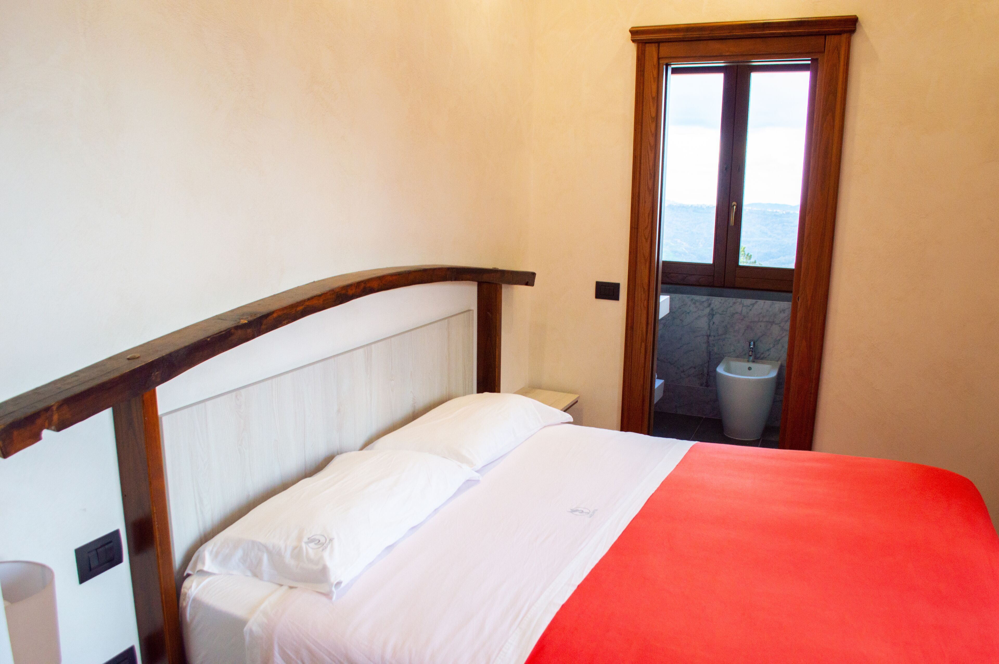Ein Schlafzimmer mit Bett, Holz-Kopfstück, roter Decke und Blick auf ein Badezimmer durch eine Tür.