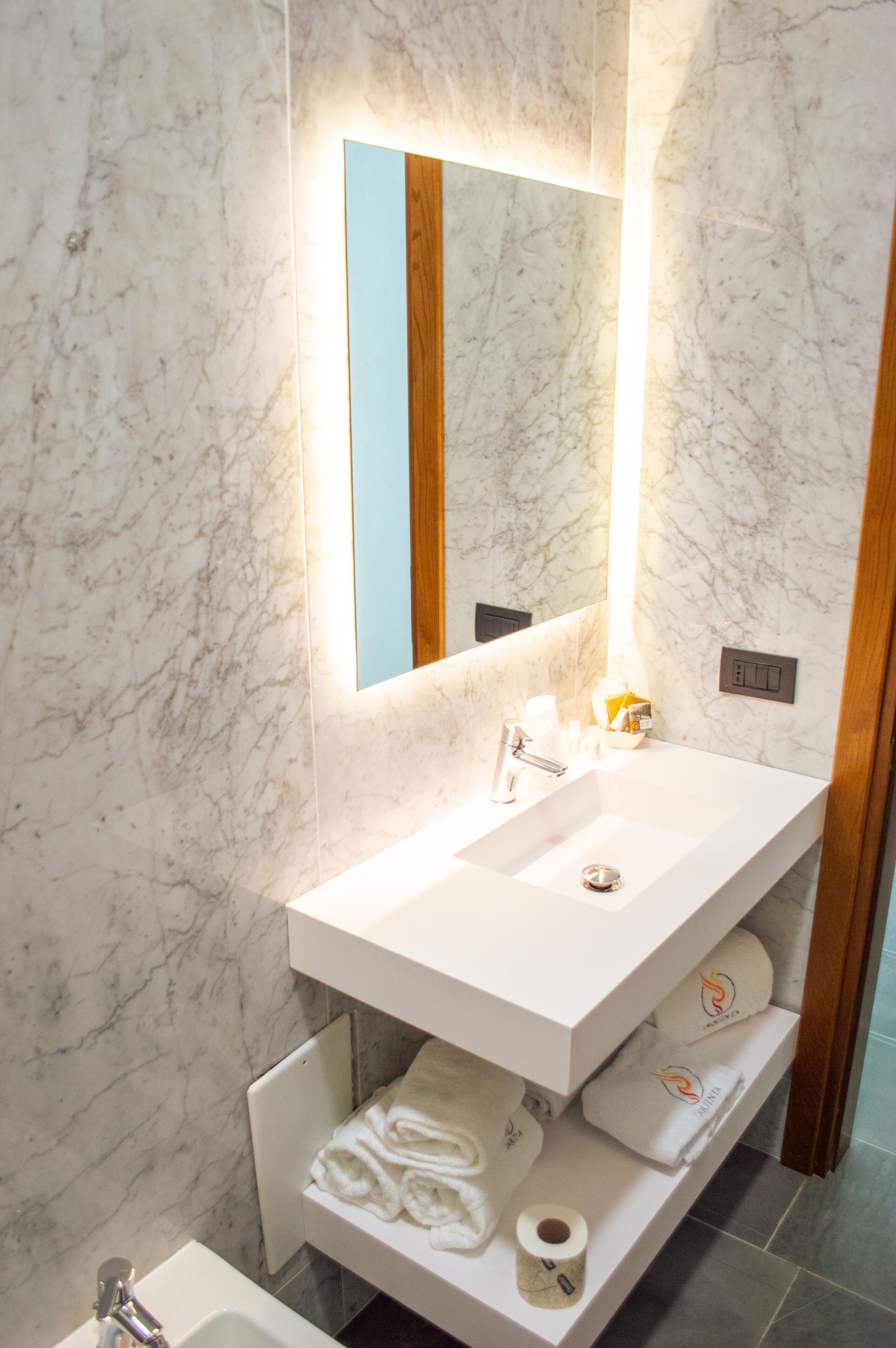 A modern bathroom with a marble wall, a white sink, a mirror, and a shelf with towels and toiletries.