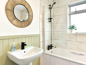 A bathroom with a white sink, a mirror with a woven frame, a shower with a black hose, and a window with a plant. - House - Sleeps up to 9 & Free Parking (Cardiff)