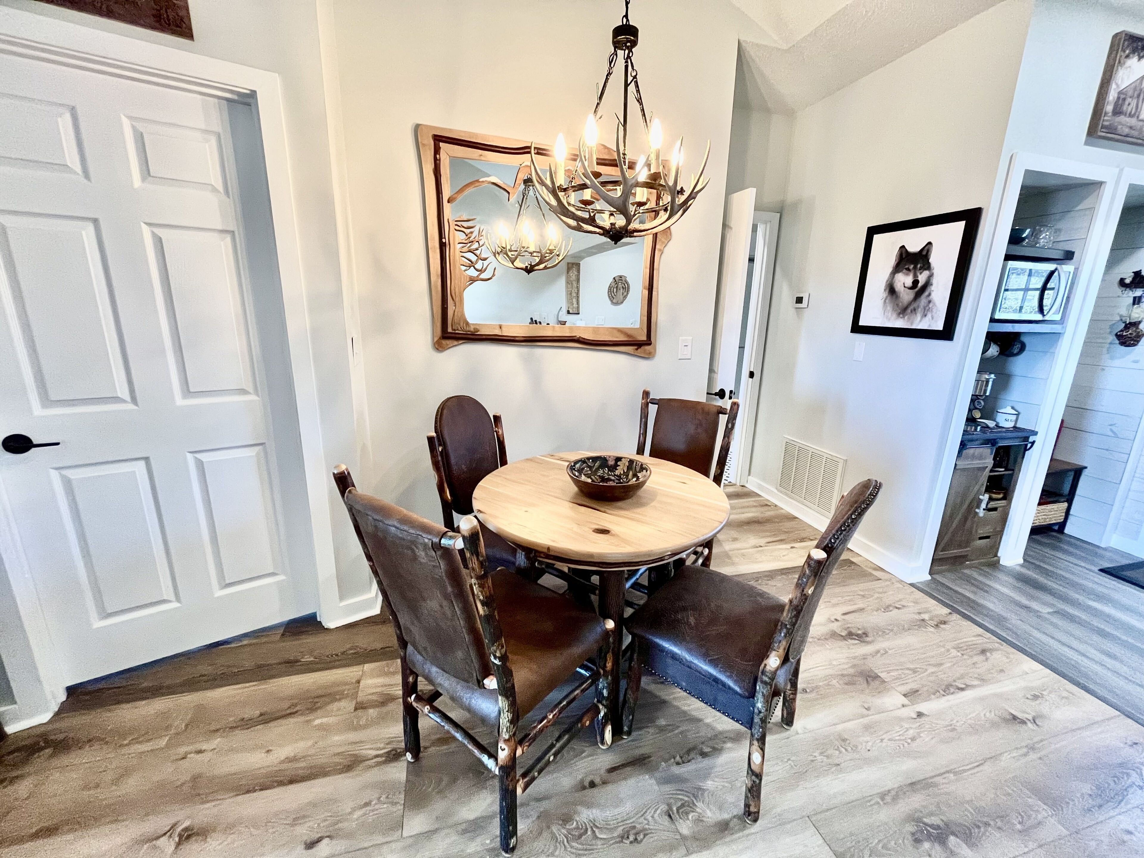 A dining room with a round table, four chairs, a chandelier, a mirror, and a framed picture on the wall.