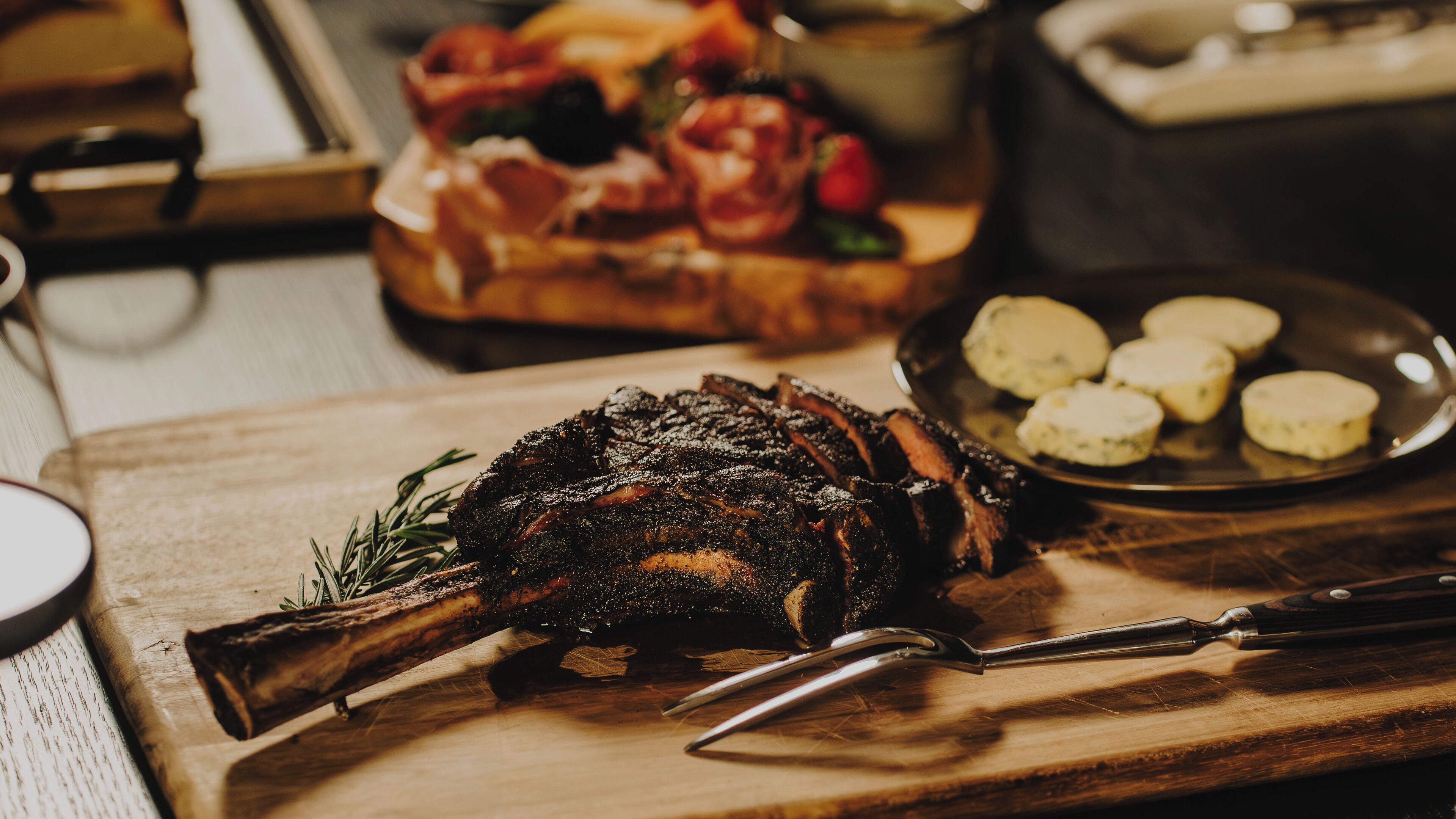 Un gigot d’agneau rôti, présenté sur une planche à découper en bois, accompagné d’un couteau et d’une assiette de petites portions de nourriture de forme ronde.