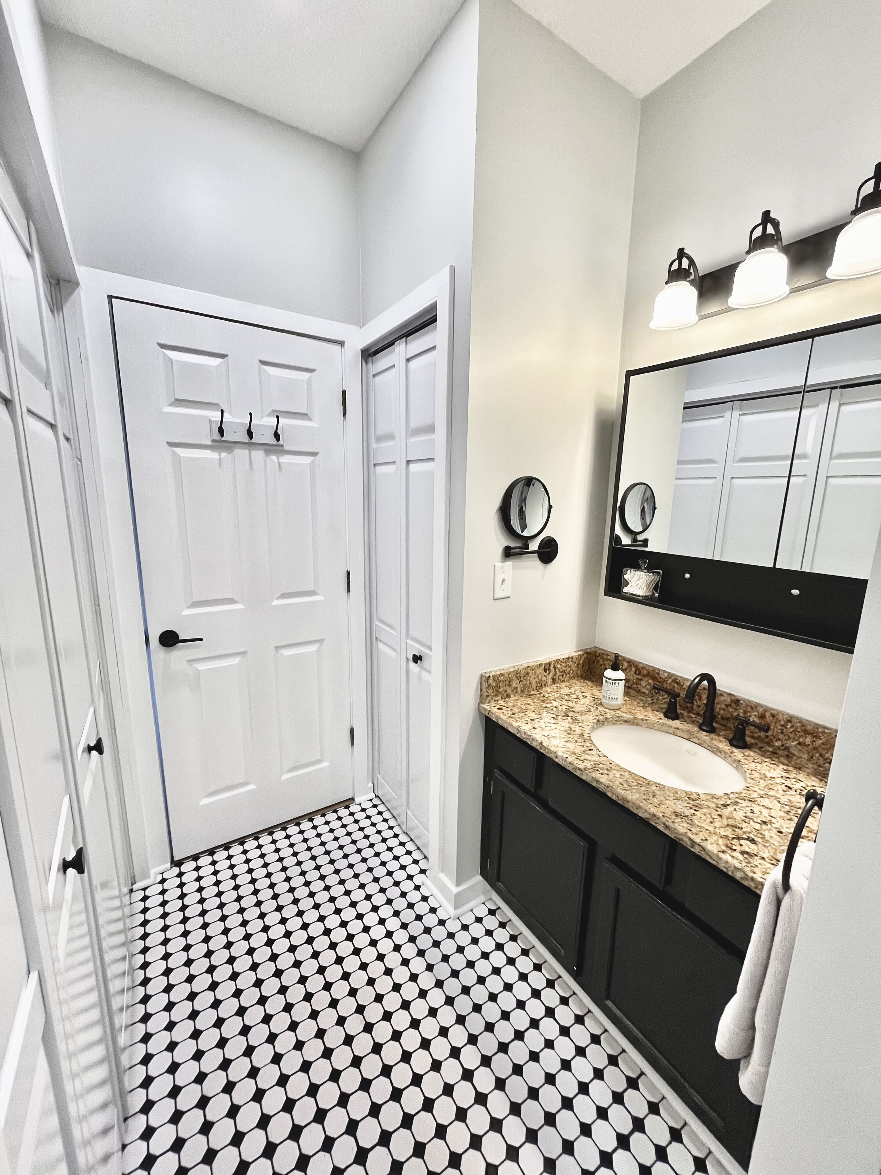 Une salle de bain avec une porte blanche, un lavabo doté d’un plan de travail en granit, un miroir et un sol à motifs noir et blanc.