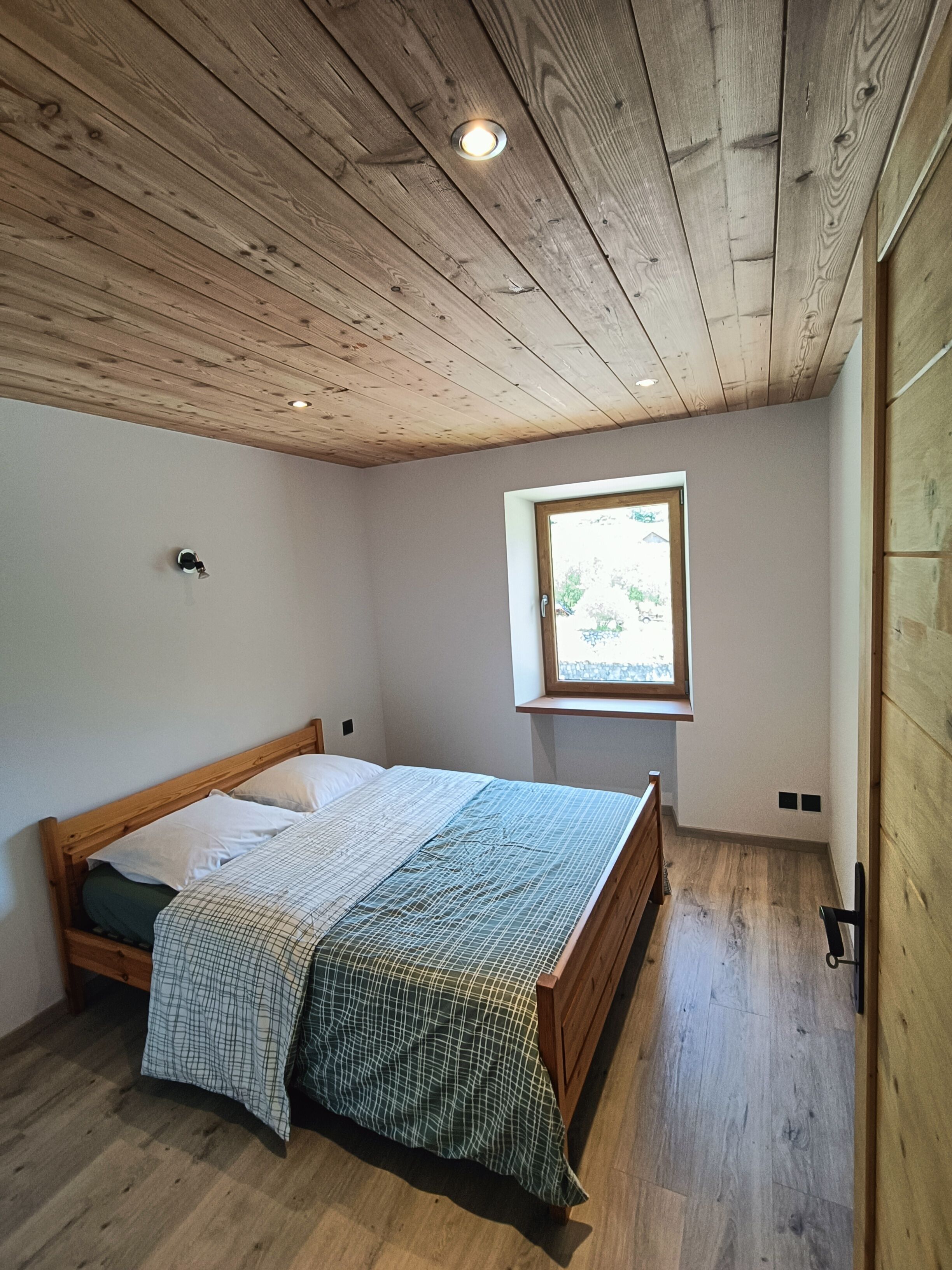 Une chambre avec un plafond en bois, une fenêtre, un lit recouvert d’une couverture à carreaux et un plancher en bois.