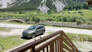 Una terraza de madera con un puente de piedra que conduce a un automóvil estacionado en un camino de grava. Al fondo, se ven montañas y un área boscosa.