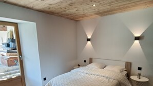 Un dormitorio con techo de madera, una cama con ropa de cama estampada, dos luces fijadas en la pared y una mesita de noche redonda y pequeña.