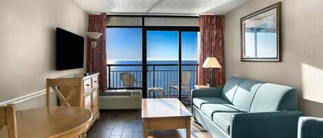 A hotel room with a balcony, a sofa, a dining table, and a TV.
