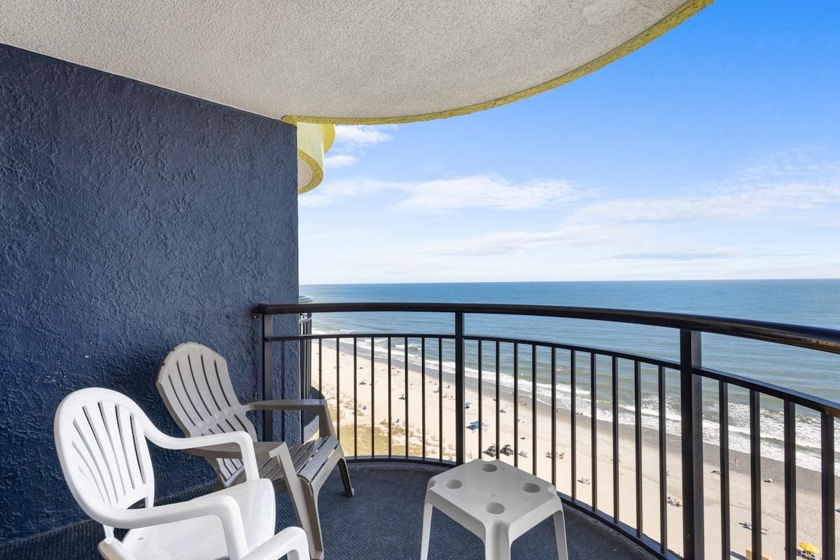 A balcony with two white chairs and a small stool, overlooking a beach with a clear view of the ocean.