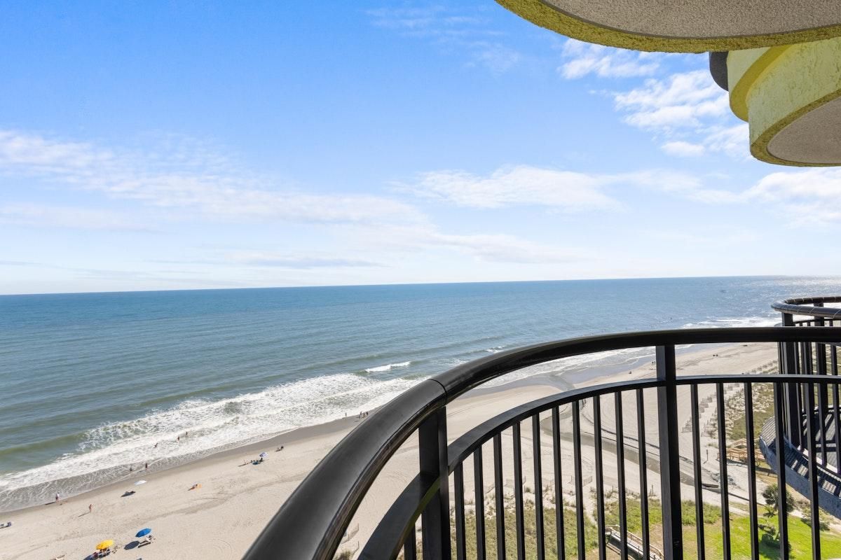A beach view from a balcony with a railing, ocean, and distant shoreline.