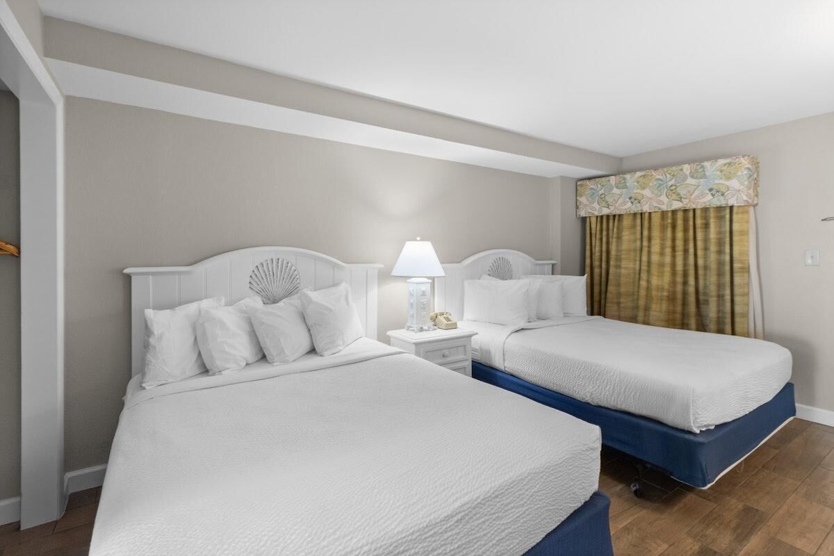 Quarto de hotel com duas camas, uma mesinha de cabeceira com abajur e uma janela com cortinas.