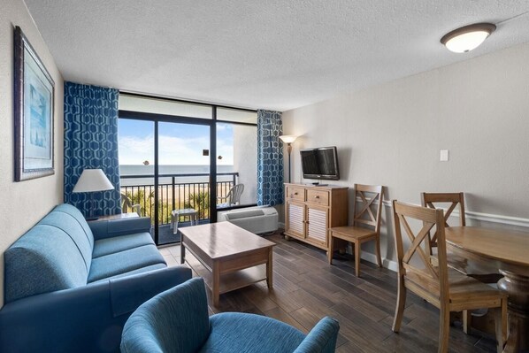 A modern living room with a sofa, a dining table, and chairs. There is a television on a stand and a window with a view of the outdoors.