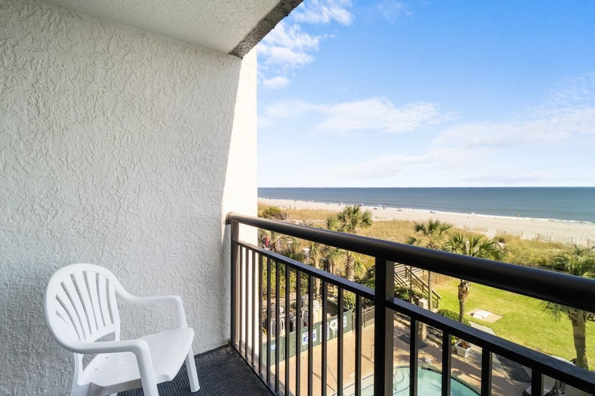 A balcony with a white chair, a pool, and a view of the beach.