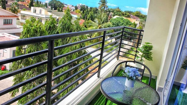 Un balcon doté d’une rambarde en métal, une petite table ronde avec un pot de fleurs, et une vue sur une ville côtière avec ses bâtiments et ses palmiers.