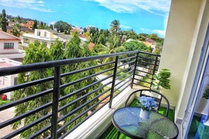 Un balcon doté d’une rambarde en métal, une petite table ronde avec un pot de fleurs, et une vue sur une ville côtière avec ses bâtiments et ses palmiers.