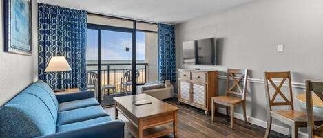 Uma sala de estar moderna com um sofá, uma mesa de centro e uma televisão. Há uma varanda com vista para o oceano.