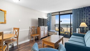A modern living room with a wooden dining table, blue armchairs, a sofa, a TV, and a balcony with patterned curtains. - Airy Oceanfront Suite: Includes Hot Breakfast (Myrtle Beach)