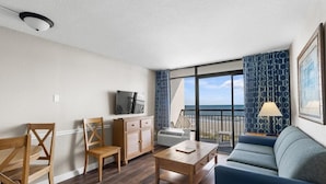 A modern hotel room with a balcony, wooden furniture, and a large window offering a view of the sea. - Majestic Oceanfront Ste 4th Flr Include Breakfast (Myrtle Beach)
