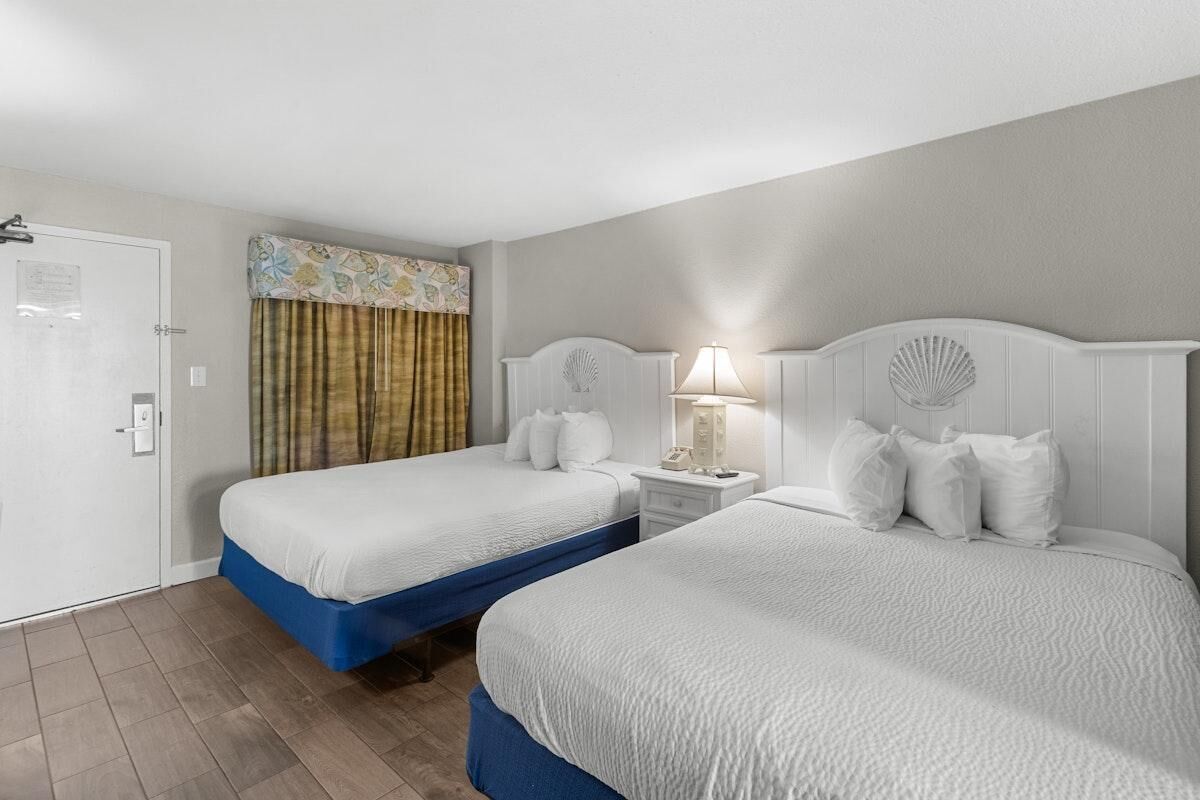 A hotel room with two beds, a door, a nightstand with a lamp, and a curtain.