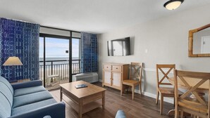 A modern living room with a sofa, coffee table, and chairs, a TV mounted on the wall, and a view of the sea through large windows. - Mesmerizing Oceanfront Ste 4th Flr w/ Breakfast (Myrtle Beach)