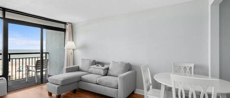 A modern living room with a grey sectional sofa, a white dining table with chairs, and a large window overlooking a balcony.