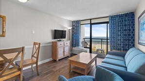A living room with a sofa, a wooden coffee table, a TV, and a balcony with patterned curtains. - Roomy Oceanfront Ste with Free Breakfast 4th Flr (Myrtle Beach)