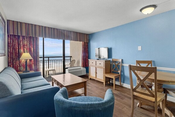 A hotel room with a balcony, a sofa, a table, a TV, and a wooden cabinet. - Spacious Oceanfront Suite: 5th Flr W Breakfast (Myrtle Beach)