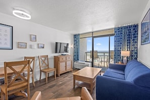 Ein Zimmer mit einem Holzespeisetisch, Stühlen, einem Sofa, einem Fernseher und einem Balkon mit gemusterten Vorhängen.