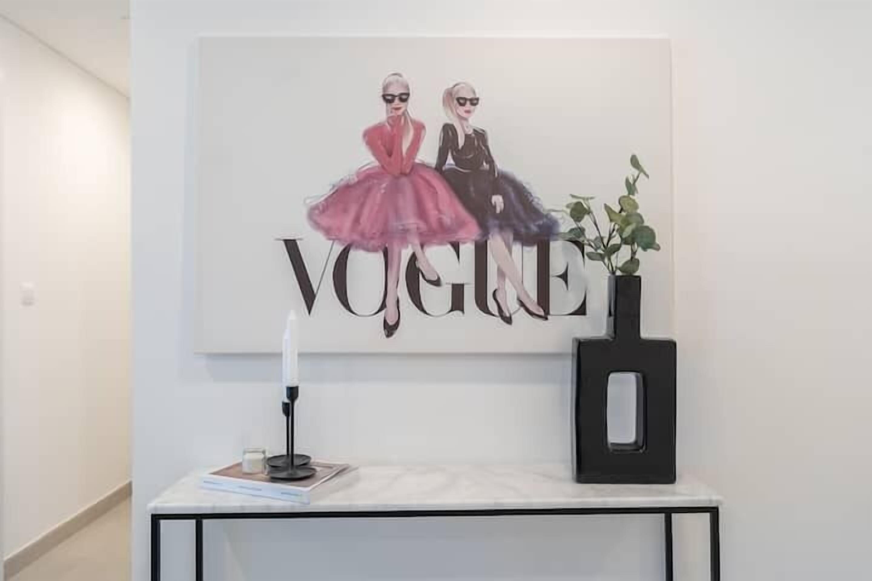 A framed artwork of two figures with the word "VOGUE" on a white wall, a black vase with a plant, and a table with a candle and a book.