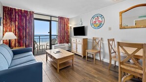 A living room with a sofa, coffee table, and chairs, a TV, a decorative plate, and a view of the sea.