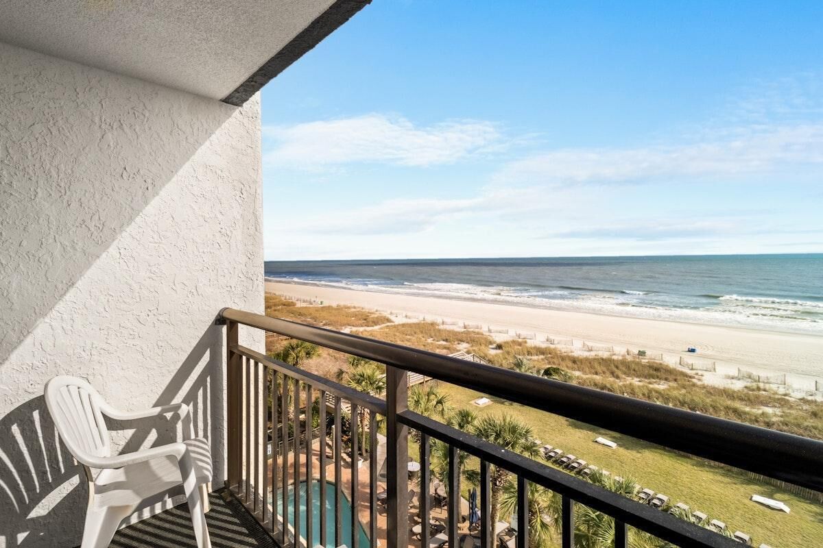 A balcony with a view of the beach, a pool, and lounge chairs.
