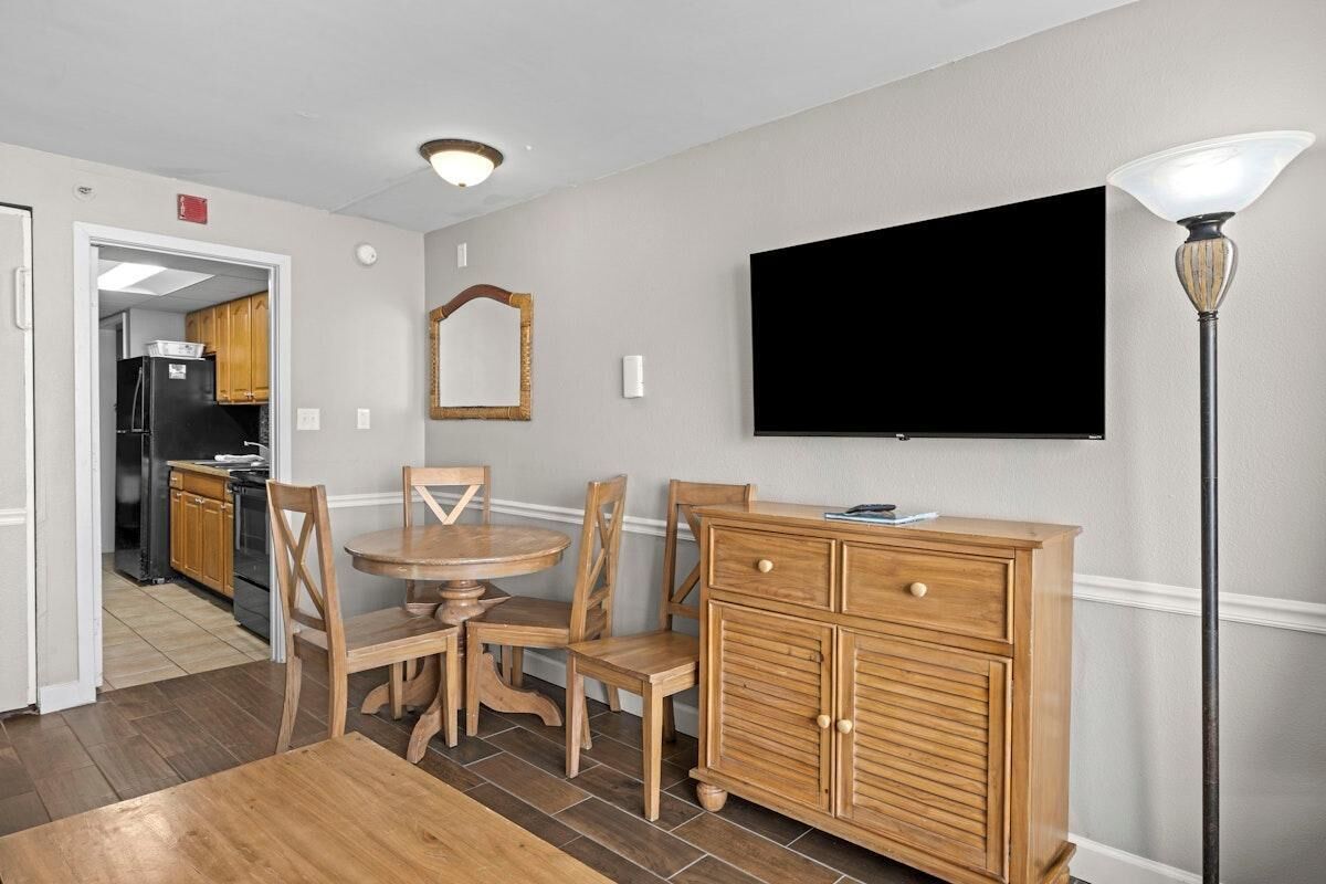 Un coin repas avec une table ronde en bois et des chaises, un meuble en bois doté de tiroirs, un téléviseur à écran plat fixé au mur et une lampe sur pied.