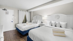 Habitación de hotel con dos camas, una puerta blanca, zona de ducha y una mesita de noche pequeña con lámpara.