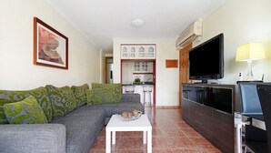 Ein Wohnzimmer mit grauem Sofa, einem weißen Couchtisch, einem Flachbildfernseher und einer Küchenzeile im Hintergrund.