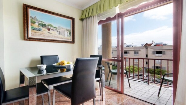 Ein Balkon mit Esstisch und Stühlen, einem Gemälde an der Wand und Blick auf Gebäude und Autos.