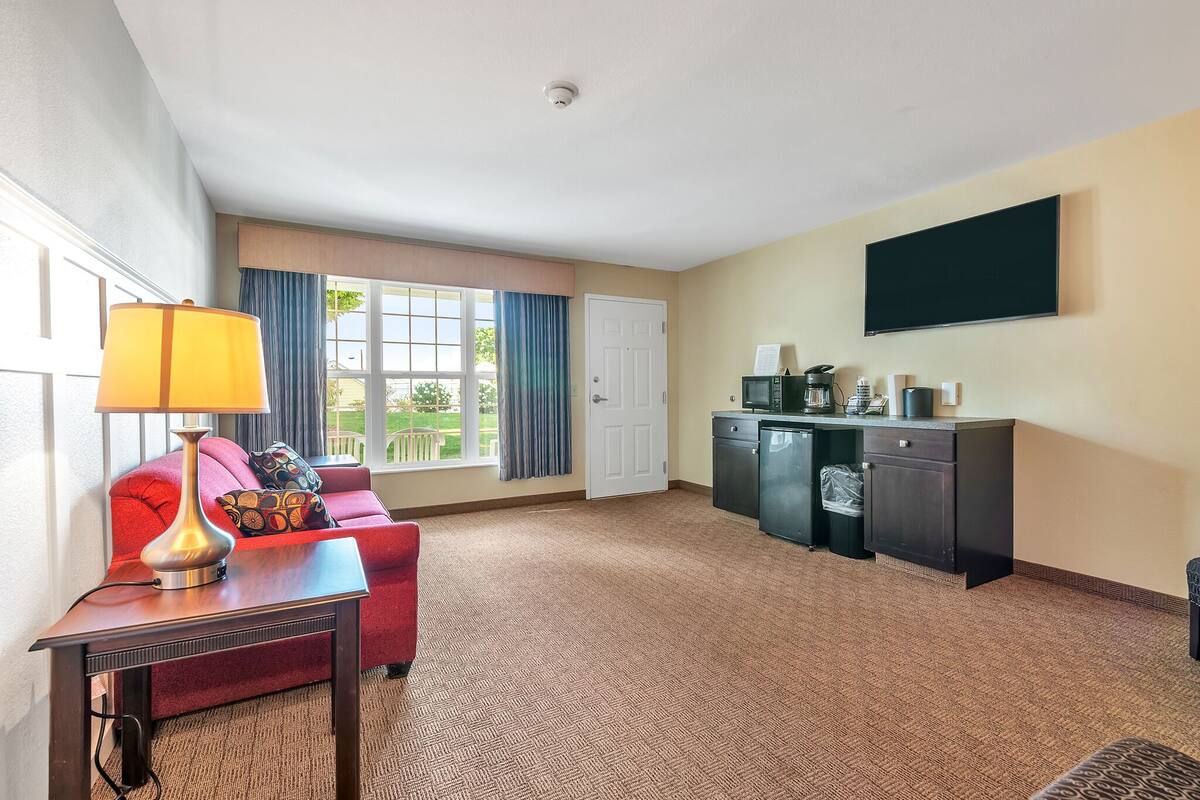 A hotel room with a red sofa, a table with a lamp, a kitchen area with a coffee maker, and a TV on the wall.