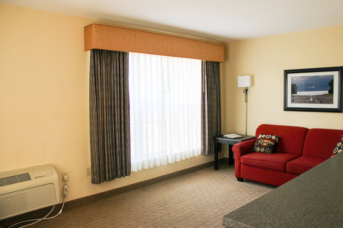 A hotel room with a red sofa, a framed picture on the wall, a window with curtains, and a table lamp.