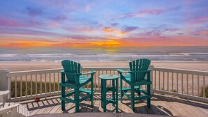 Une terrasse en bois avec deux fauteuils Adirondack verts et une petite table, donnant sur une plage où l’on peut admirer le coucher de soleil.