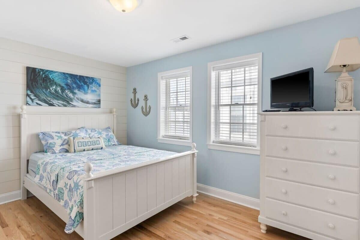 A bedroom with a white bed, a dresser, two windows with blinds, a painting on the wall, and a television on a stand.
